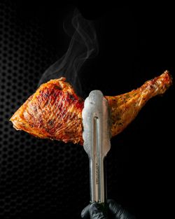 Morceau de poulet grillé tenu avec une pince métallique, dégageant de la vapeur sur fond noir texturé.