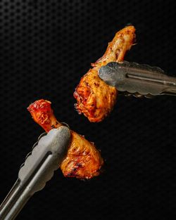 Deux morceaux de poulet grillé saisis avec des pinces métalliques sur fond noir texturé, cuisson dorée et épicée.
