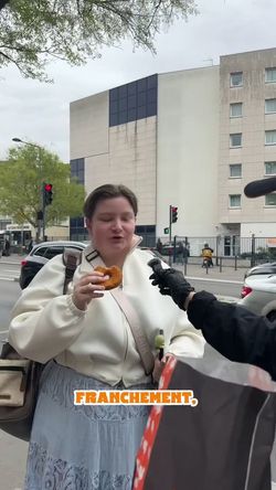 Une personne tenant un sandwich, devant un bâtiment urbain, prête à recevoir un autre objet d'une main gantée.