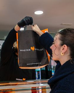 Un sac à emporter noir et orange avec le logo MASTER POULET remis à une cliente dans un restaurant.