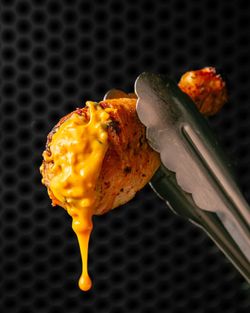 Poulet grillé en rôtisserie avec sauce jaune fondante, tenu par une pince métallique sur fond noir à motifs hexagonaux.