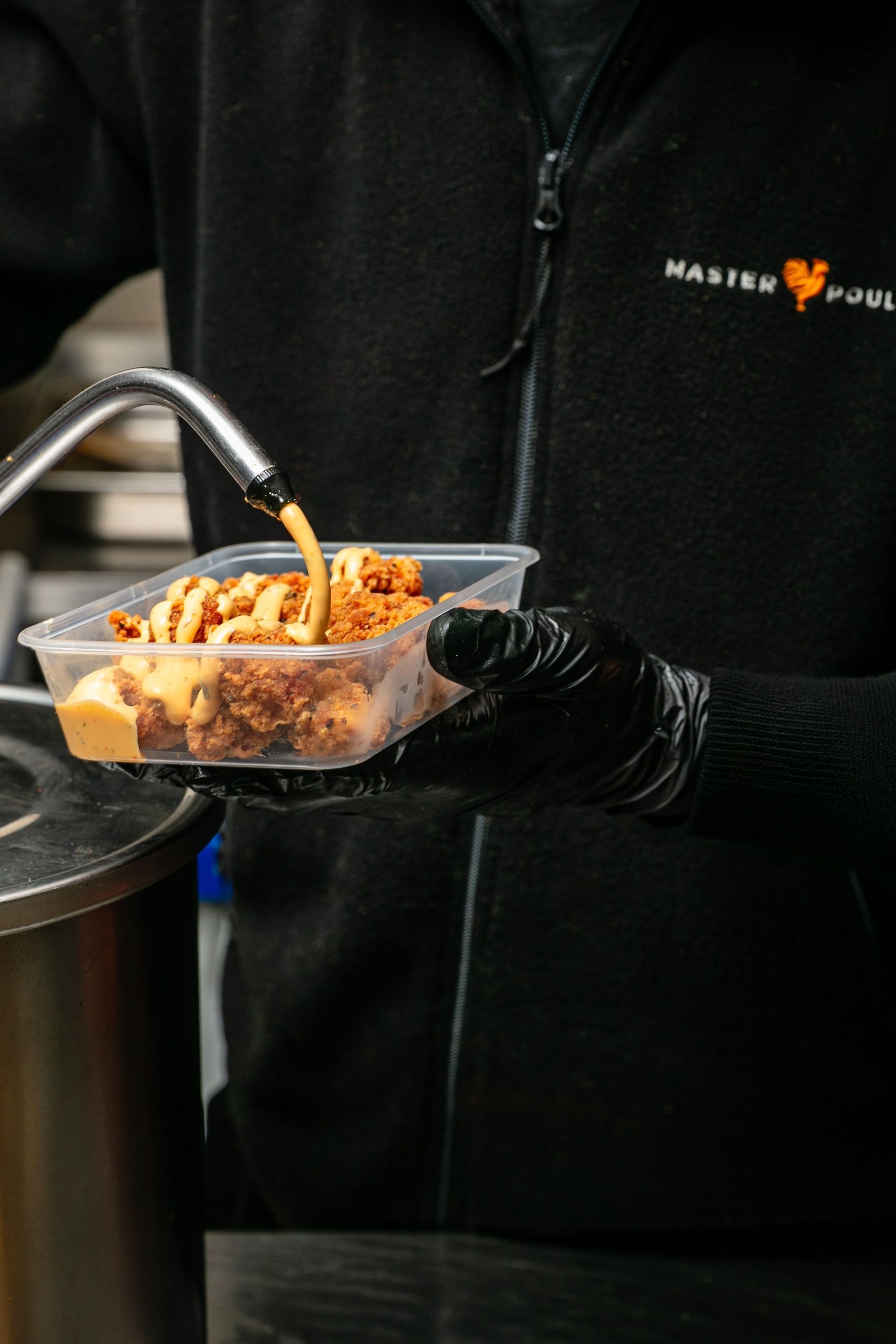 Un employé de MASTER POULET verse une sauce sur du poulet frit dans une barquette plastique à emporter.