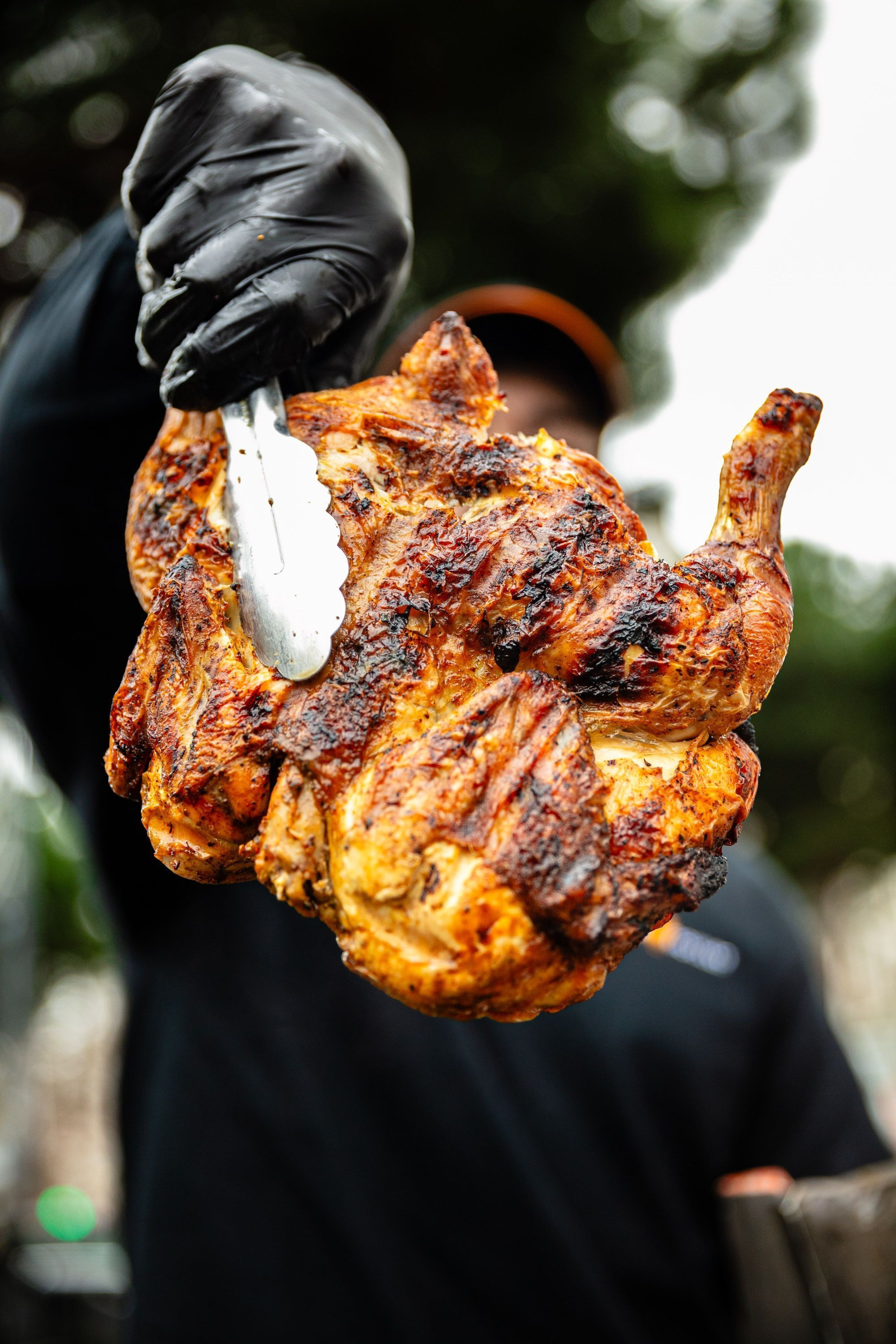 Poulet rôti grillé tenu avec une pince par une main gantée noire, cuisson bien dorée et croustillante visible